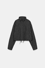 Cazadora negra con estampado de lunares blancos, cuello subido, manga larga, bolsillos delanteros, bajo ajustable con cordones y cierre frontal con cremallera y solapa con botones ocultos.