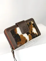 Billetera grande tipo sobre con correa de mano, confeccionada en cuero marrón con detalle central de cuero con pelo vacuno en tonos marrón, negro y blanco. Presenta cierre perimetral y solapa frontal con broche magnético y logo de búho grabado.