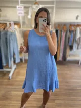 Vestido corto sin mangas, color gris topo, con escote redondo y detalle de volado en el ruedo. El tejido presenta una textura arrugada característica del lino.