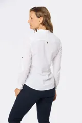 Camisa blanca de manga larga con cuello y tapeta de medio botón. Presenta dos bolsillos frontales con tapa y botones metálicos. Las mangas tienen presillas para sujetar el puño enrollado.