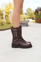 Bota corta de cuero marrón oscuro, estilo motociclista, con caña media arrugada y tres hebillas ajustables en el lateral. Posee una tira decorativa con argolla metálica alrededor del tobillo y suela track gruesa.