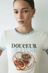Remera corta de algodón blanca con ribetes celestes en el cuello y las mangas. Presenta un estampado frontal con la inscripción "Douceur de vivre" y una ilustración de tostadas con mermelada y un frasco.