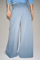 Pantalón wide leg de tiro medio confeccionado en hilatura de algodón, color beige, con trabillas en la cintura, bolsillos laterales ocultos y detalle de pliegues en el delantero. Cierre frontal con cremallera, botón y ganchos metálicos.