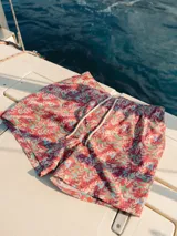 Short de baño para hombre con estampado de hojas en tonos verde, celeste y blanco sobre fondo rosado. Cuenta con elástico en la cintura, cordón ajustable y tres bolsillos.