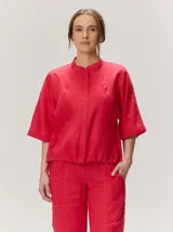 Chaqueta de lino y rayón color rojo, con cuello mao, mangas cortas holgadas y cierre frontal oculto. Presenta pespuntes decorativos a contratono.