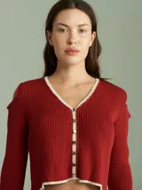 Saco de punto acanalado color bordó con detalles en color crudo en el cuello, plaqueta de botones y ruedo. Presenta aberturas en los hombros y cierre frontal con botones pequeños.