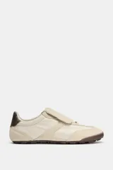 Championes deportivos de estilo minimalista en color beige, confeccionados en una combinación de materiales textiles y gamuza. Presentan una lengüeta superpuesta con cierre de gancho, punta redonda y un detalle de textura oscura en el talón. Suela plana de goma en tono marrón.