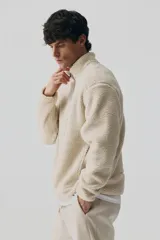 Campera deportiva de tejido sherpa color beige, con cuello elevado, cierre frontal completo, mangas raglán, bolsillo superior con cierre y bolsillos laterales. Presenta terminaciones elásticas en puños y ruedo.