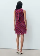 Vestido corto sin mangas con cuello alto, confeccionado en tejido estampado con patrón geométrico en tonos rojos y violetas.