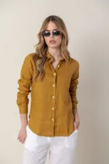 Camisa de lino color mostaza con cuello clásico, cierre frontal de botones y mangas largas con puños abotonados. Presenta un pequeño bordado distintivo en el ruedo inferior.