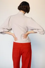 Blusa de cuello subido con escote en pico y cierre frontal con botones forrados. Presenta mangas abullonadas de largo tres cuartos con elástico en los puños y detalle de lazos en el cuello y el frente.