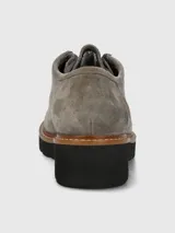 Zapato estilo mocasín o derby de mujer, color gris, confeccionado en gamuza. Presenta suela plataforma negra con borde de cuero marrón cosido y cordones grises que pasan por ojales metálicos dorados.