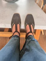 Ballerinas de gamuza color marrón oscuro con diseño tipo T-strap y detalles de costuras decorativas en color verde oliva. Cuentan con cierre de pulsera al tobillo y suela plana.