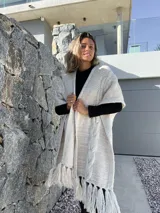 Ruana tejida en lana merino, color beige y crudo, con diseño de trenzas verticales en el frente y terminación con flecos en el ruedo.