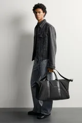 Bolso de viaje tipo bowling fabricado en combinación de nylon y detalles en cuero sintético, de color negro. Cuenta con compartimento principal con cierre de cremallera, bolsillo frontal plano, dos asas de mano y un asa de hombro ajustable y extraíble.