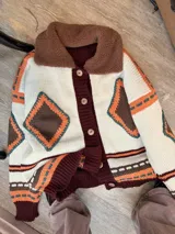 Campera tejida de punto con diseño geométrico en tonos crema, marrón y naranja. Presenta un cuello de corderito en color marrón, cierre frontal con botones y puños y ruedo acanalados en color bordó.