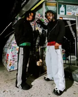 Pantalón deportivo negro de corte ancho, con tres franjas blancas verticales a lo largo de las piernas. La cintura es elástica y presenta un forro interior con estampado a cuadros en tonos verde y negro.