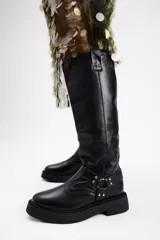 Bota de caña alta confeccionada en cuero vacuno color negro, con suela track de plataforma y detalle de tira con argolla metálica y tachas en el tobillo.