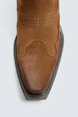 Bota estilo cowboy de cuero color marrón con acabado serraje engrasado, caña media con pespuntes decorativos, tiradores laterales, tacón ancho y punta cuadrada.