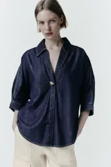 Camisa de denim azul oscuro con corte amplio, cuello solapa, escote en pico y mangas tres cuartos. Presenta un aplique metálico dorado en el centro y bajo asimétrico.