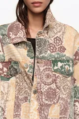 Chaqueta larga de corte oversize confeccionada en algodón con estampado patchwork en tonos beige, verde y marrón. Cuenta con cierre frontal con botones, cuello camisero, mangas largas, bolsillos con solapa en el pecho y bolsillos laterales.