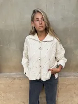 Saco tejido de punto con diseño de ochos y trenzas, color crudo. Presenta cuello camisero, cierre frontal con botones joya y mangas holgadas con puños acanalados.