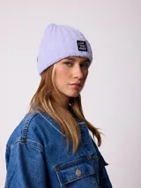 Gorro tipo beanie de punto acanalado en color lila, con parche rectangular negro en el frente que incluye la inscripción 'EAT THE WAVE'.