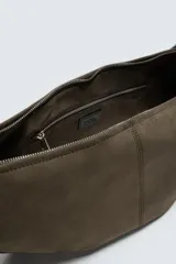 Bolso de hombro con diseño de media luna, confeccionado en gamuza color marrón. Cuenta con un compartimento principal con cierre de cremallera y un bolsillo interior con cierre. Posee un asa de hombro integrada.