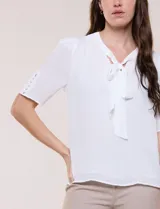 Blusa blanca de corte recto y tejido liviano de gasa con forro interno delantero. Presenta escote en V con cuello redondo y lazo central para anudar, mangas cortas con hilera de botones forrados en los laterales y ruedo ligeramente redondeado.