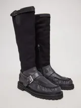 Bota híbrida de caña alta confeccionada en lona negra en la parte superior y cuero negro en la base tipo mocasín, con plataforma, suela track y tira con hebilla metálica cruzada en el empeine.