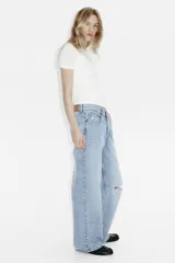 Jean de corte wide leg con diseño de cinco bolsillos, confeccionado en denim de algodón rígido. Presenta un lavado claro con efecto desgastado y un detalle de rotura en la rodilla izquierda. Cuenta con cintura clásica, cierre de botón y bragueta.