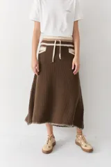 Falda midi de lana en color marrón, con diseño acampanado y tejido acanalado. Presenta una cintura elástica con cordón de ajuste, detalles de rayas en tonos beige y blanco en la parte superior, y costuras decorativas en el ruedo.