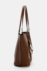Bolso tipo shopper de color marrón, con diseño minimalista, doble asa de hombro y cierre magnético. Incluye un colgante decorativo con detalle de cinta en tonos claros y un bolsillo interior amplio.