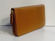 Billetera rectangular de cuero color beige con cierre perimetral y tirador de cuero. Cuenta con múltiples compartimentos internos para tarjetas, documentos y billetes, además de un bolsillo interno con cierre para monedas y forro textil en color rojo.