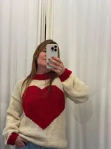 Buzo tejido a mano en lana merino, de color crema con un gran corazón rojo en el centro y detalles en rojo en el cuello y los puños. Presenta un diseño de manga larga y corte holgado.