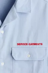 Camisa de manga corta color celeste, de corte relajado, con cuello abierto, bolsillos delanteros con solapa y cierre de botones. Cuenta con detalle de texto bordado en rojo sobre el bolsillo superior.