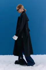 Trench midi de color negro, confeccionado en una mezcla de algodón y nylon con propiedades repelentes al agua. Presenta un diseño holgado con tajos laterales, bolsillos internos y detalle de etiqueta en la espalda.