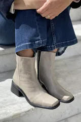 Botas cortas color visón de cuero charolado, con punta cuadrada, caña media y detalle de arnés con hebillas plateadas. Taco cuadrado de altura media color negro.