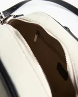 Cartera tipo baúl bicolor, combinando cuerpo de lona beige con detalles de símil cuero negro en las asas, bordes y correa. Cuenta con doble asa de mano, cierre superior y correa ajustable y desmontable para usar al hombro o cruzada.