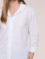 Camisa de silueta holgada y largo extendido, confeccionada en poplín elástico de color blanco. Presenta cuello solapa abierto en V con cartera frontal de abotonadura oculta, hombros caídos y mangas tres cuartos. Cuenta con espalda con canesú, pliegue central y ruedo recto con pequeñas aberturas laterales.