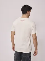 Remera color crema de hombre, marca Rusty, con estampado frontal en color rojo con el logo de la marca y la leyenda "Rusty Worldwide Supply Co. Since 1985".