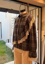 Camisa de franela a cuadros en tonos marrones y grises, con diseño de volados cruzados en el frente adornados con encaje crudo, mangas largas con puños acampanados y cierre frontal de botones.