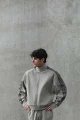 Campera deportiva de color gris topo, con cuello alto, cierre frontal bidireccional y bolsillos laterales. Presenta franjas laterales negras en las mangas y puños y bajo de punto acanalado.
