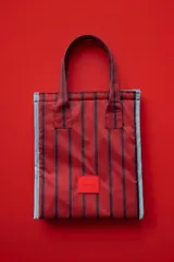 Lunchera rectangular con diseño de rayas verticales en tonos rojos y bordes en color celeste. Cuenta con doble asa superior y parche frontal con el logo de la marca.