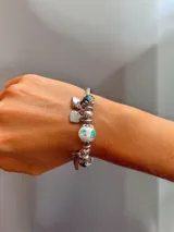 Pulsera rígida plateada con dijes colgantes y cuentas decorativas. Incluye cuentas de piedra veteada, cristales facetados en tono verde petróleo, una cuenta de cerámica blanca con diseño floral y múltiples dijes de corazón, uno de ellos con la frase 'you love you' grabada.