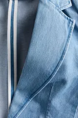 Blazer de denim de algodón celeste, con solapas de muesca, hombreras y mangas tres cuartos fruncidas. Cuenta con bolsillos delanteros con solapa y diseño abierto sin cierre.