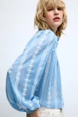 Blusa corta de color blanco con diseño de bordados perforados, cuello solapa, cierre frontal con botones y mangas largas abullonadas.