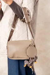 Cartera bandolera color chocolate de diseño compacto, con correa ajustable de tela y detalle de monedero circular y dije de perro salchicha colgante.