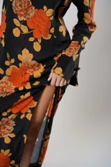 Vestido largo negro con estampado de flores naranjas y amarillas, cuello mao con abertura en forma de gota, mangas largas acampanadas con botones en los puños, tajo lateral y flecos en el ruedo.