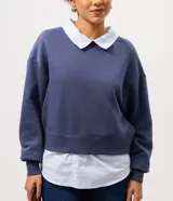 Buzo azul de corte amplio confeccionado en mezcla de algodón y poliéster, con diseño superpuesto que incluye cuello clásico y dobladillo en tono contrastante simulando una camisa por debajo.